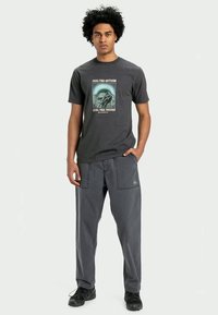T-shirt cinza com gráfico de montanha e texto, combinada com calças cargo cinza escuras com bolsos e detalhes de logótipo, usadas com sapatos pretos.