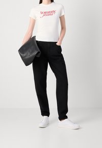 Femme portant un t-shirt blanc "Tommy Jeans", pantalon noir, baskets blanches, tenant une veste en cuir noire pliée sur un fond gris uni.