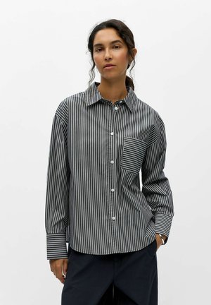 Femme aux cheveux foncés portant une chemise boutonnée noire et blanche à rayures verticales avec une poche poitrine rayée contrastante.