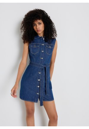 Donna con capelli ricci che indossa un vestito di jeans senza maniche con bottoni frontali e cintura in vita, in piedi contro un muro chiaro e semplice.