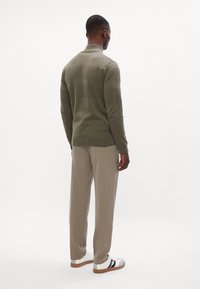 Pullover lavorato a maglia verde oliva con motivo a strisce testurizzate e colletto alto, abbinato a pantaloni marrone chiaro e sneakers bianche con dettagli neri.