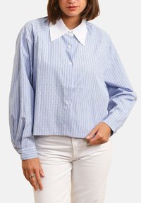 Chemise rayée bleue avec un col blanc, présentant des lignes verticales et de petits points. Coupe décontractée avec des manches longues et une fermeture à boutons.
