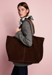 Brown suede tote bag with a rectangular shape, featuring a stitched design and two shoulder straps. Worn with a houndstooth coat against a pink background.