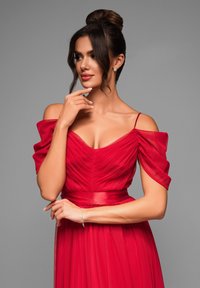 Robe de soirée rouge avec un corsage ajusté, mettant en avant des plis et des manches dénudées. La jupe tombe doucement, resserrée à la taille par une ceinture.
