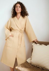 Beige waffle-textúrájú köntös hosszú ujjakkal és megkötő övvel, két elülső zsebbel. Egy csíkos párna fodros szélekkel fekszik mellette.
