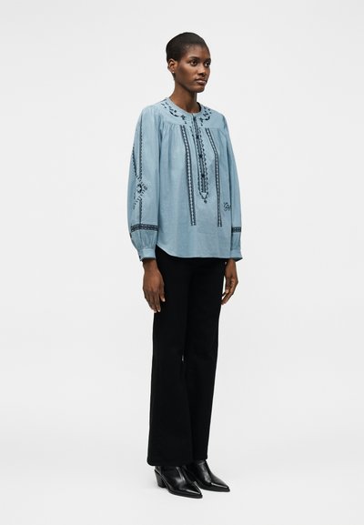 Γυναίκα όρθια, φορεμένη με ανοιχτό μπλε κεντημένη μπλούζα με μακρύ μανίκι, μαύρο παντελόνι και μαύρα μυτερά αστραγάλια, σε λευκό φόντο.