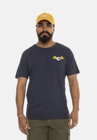 Hombre con barba y pendientes en el tabique, llevando una gorra amarilla, una camiseta de color azul marino con un gráfico de un ojo alado, y pantalones cortos de carga en color oliva, de pie contra un fondo blanco.