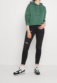 Osoba oblečená v zelené zkrácené mikině s kapucí, černých roztrhaných skinny džínech, bílých ponožkách a černobílých teniskách New Balance stojí na jednobarevném pozadí.