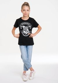 Black short-sleeve t-shirt featuring a white Hogwarts crest graphic, paired with light blue cuffed jeans and white sneakers with pink accents.