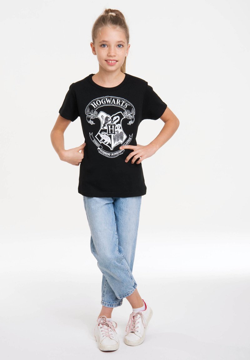 Black short-sleeve t-shirt featuring a white Hogwarts crest graphic, paired with light blue cuffed jeans and white sneakers with pink accents.
