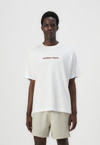 Camiseta de algodón blanca con cuello redondo y mangas cortas. En la parte delantera presenta "atelier dept." en una fuente serif roja. El modelo lleva pantalones cortos beiges.