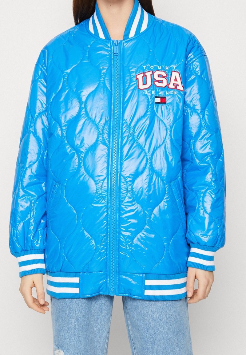 Personne portant une veste bomber matelassée bleu vif avec des poignets, un col et un ourlet rayés blancs, ainsi qu'un logo USA sur le côté gauche de la poitrine.