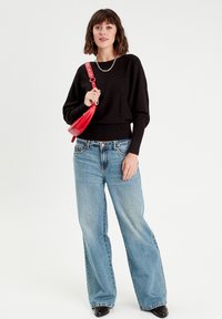 Pull en maille noir avec un ourlet côtelé, associé à un jean large bleu clair. Le modèle tient un sac à main rouge avec une bandoulière. Bottes noires.
