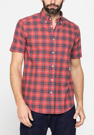 Uomo che indossa una camicia a maniche corte a quadri rossa e blu navy con jeans scuri, in piedi davanti a uno sfondo bianco uniforme.