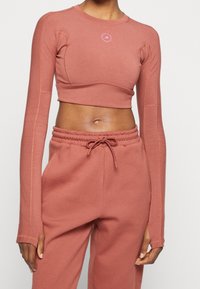 Nainen, jolla on päällä pölyruusunvärinen pitkähihaisuus crop-top ja siihen sopivat korkeavyötäröiset collegehousut, joiden naru on solmittu eteen.