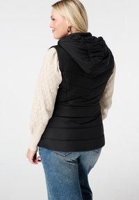 Zwarte gewatteerde vest met capuchon, met een gestikte ontwerp. Draag boven een crèmekleurige textuur long-sleeve top, gecombineerd met blauwe spijkerbroek.