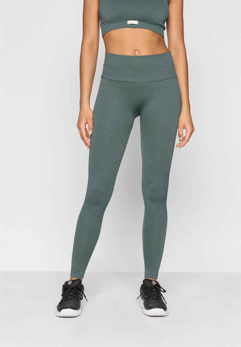 Conjunto de athleisure verde com nervuras, composto por um top cropped e leggings de cintura alta. O material parece elástico. Usado com ténis de desporto pretos.