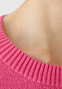 Pull rose tricoté avec un col côtelé ; le tissu texturé présente un motif en losange près du col, se fondant avec une teinte de peau douce.