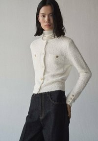 Weißer strukturierter Pullover mit hohem Kragen, Knopfverschluss und zwei Brusttaschen. Kombiniert mit dunklen, hochgeschnittenen Jeans.