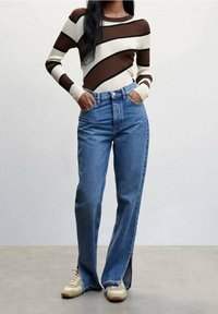 Henkilö, jolla on päällään raidallinen ruskea, kerma ja musta neulepaita, siniset korkeavyötäröiset farkut, joissa etummaiset halkiot, ja beiget lenkkarit, seisomassa käsien ollessa taskuissa.