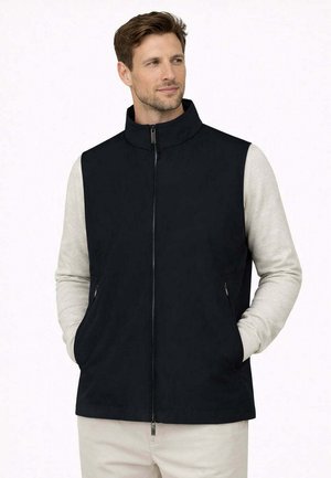 Uomo che indossa un gilet nero senza maniche con cerniera sopra una camicia a maniche lunghe grigio chiaro, con le mani in tasca, che guarda leggermente verso destra.