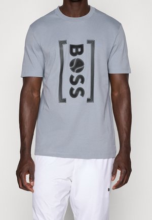 Homme portant un t-shirt gris clair à manches courtes avec texte noir "BOSS" et graphique balle de tennis, associé à un pantalon blanc.