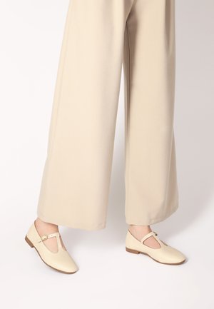 Beige Leder Schuhe mit T-Riemen-Design, flachen Sohlen und runder Zehenpartie. Die Schuhe haben eine kleine Schnallenakzentuierung.