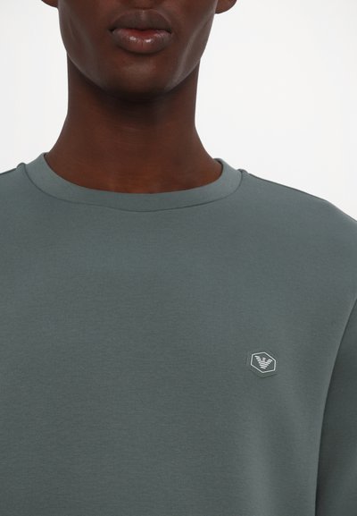 Sudadera de cuello redondo gris verdoso, fabricada en un tejido suave, con un pequeño logo hexagonal en un tono más claro en la parte izquierda del pecho.