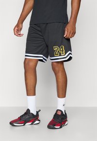 Pantalones cortos deportivos negros con estampado amarillo "24", rayas blancas en los dobladillos, combinados con zapatillas atléticas negras y rojas y calcetines blancos.