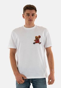Camiseta de algodón blanca con un gráfico de un oso vestido con un uniforme deportivo rojo y un balón de baloncesto, situado en la zona del pecho izquierdo. Mangas cortas.