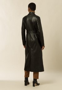Černý kožený dlouhý kabát s páskem v oblasti pasu, přiléhavý střih a jednoduchý design. Nošený s hnědými kalhotami a černými botami na podpatku.