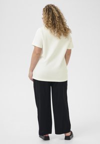 Off-white t-shirt met korte mouwen, gecombineerd met losse zwarte broeken en stevige zwarte schoenen, tegen een plain lichte achtergrond.