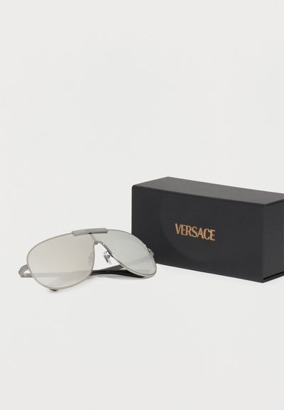 Γυαλιά ηλίου Versace με ασημί απόχρωση, μονός κυρτός φακός και ρυθμιζόμενα μαξιλαράκια μύτης, δίπλα σε μαύρο κουτί με χρυσό λογότυπο Versace.