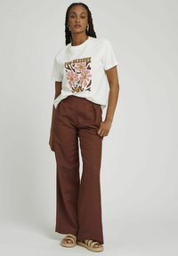 T-shirt en coton blanc avec un motif floral en rose et marron, associé à un pantalon large à taille haute couleur rouille et des sandales tressées.