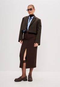Blazer corto marrone sopra una camicia azzurro chiaro, abbinato a una gonna midi marrone con uno spacco laterale e mocassini marroni. Occhiali da sole chiari e rettangolari.