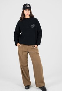 Junge Frau, die eine schwarze Mütze und einen Pullover mit Logo, braune weite Hosen und schwarze Schuhe trägt, mit den Händen in den Taschen vor einem einfarbigen Hintergrund steht.