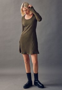 Olivgrünes, geripptes Kleid mit Rundhalsausschnitt, langen Ärmeln und seitlichen Schlitzen, kombiniert mit schwarzen klobigen Stiefeletten.