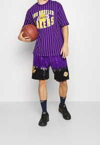 Un joven sostiene una pelota de baloncesto, vistiendo una camiseta a rayas morada de Los Angeles Lakers y pantalones cortos de tie-dye. Zapatillas negras y calcetines azul marino completan el look.