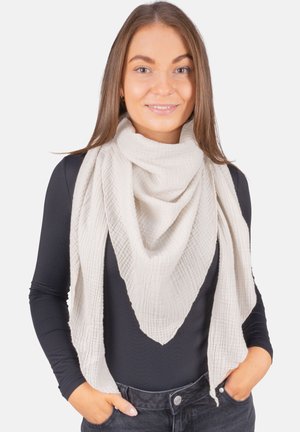 Femme aux cheveux bruns lisses portant une écharpe crème texturée sur un chemisier noir à manches longues et un jean foncé, les mains dans les poches, souriant légèrement.