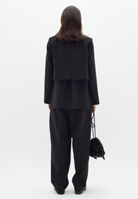 Chaqueta negra estructurada con un diseño corto, superpuesta a una blusa larga negra y pantalones holgados; sosteniendo un pequeño bolso negro texturizado.