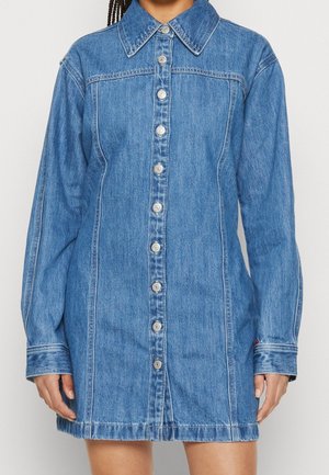 Blauwe denim jurk met lange mouwen, knoopsluiting aan de voorkant, kraag en zichtbare stiksels gedragen door een persoon die voor een lichte achtergrond staat.