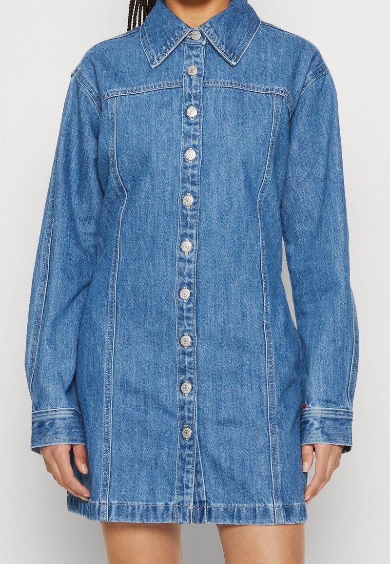 Robe en denim bleu à manches longues avec fermeture boutonnée sur le devant, col et coutures visibles portée par une personne debout devant un fond clair.