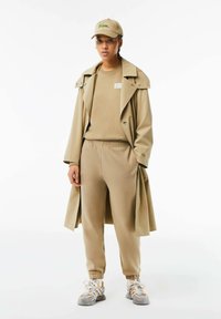 Beige trenchcoat över en matchande sweatshirt och joggers. Innehåller en Lacoste-logga cap och ljusgrå sneakers med gula accenter.