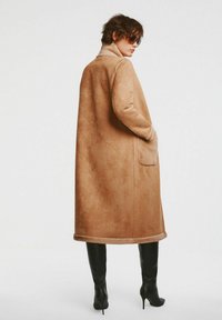Karmelowy zamszowy płaszcz trenchtowy o prostym kroju, z dużym kołnierzem, bocznymi kieszeniami i miękką teksturą. Noszony z czarnymi, wysokimi na uda botkami na obcasie.