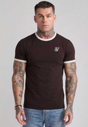 Mand med tatoveringer på nakke og arme iført en mørkebrun tætsiddende t-shirt med hvid kant og lyseblå jeans, stående mod en ensfarvet baggrund.