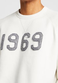 Gros plan sur une personne portant un sweat-shirt blanc avec de grands chiffres gris texturés "1969" sur la poitrine.
