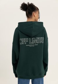 Green oversized hoodie with a drawstring hood, featuring white text "OFF LIMITS DANGEROUS AREA BERLIN 2001" on the back. Soft texture.