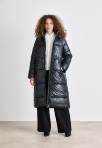 Casaco puffer preto com uma textura brilhante, de comprimento longo, fecho de zip, dois bolsos frontais com detalhes em dourado e um colarinho alto.