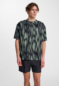 Grafisk t-shirt med ett flytande svart och grönt abstrakt mönster, rundad halsringning, korta ärmar och en liten vit chevron-logotyp på bröstet.