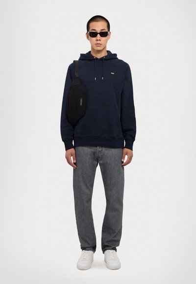 Jūras spēku krāsas kapucis, pelēki džinsi, baltas kedas un melna pleca soma. Modelis valkā melnus saulesbrilles. Vienkārša, ikdienišķa apģērba kombinācija.